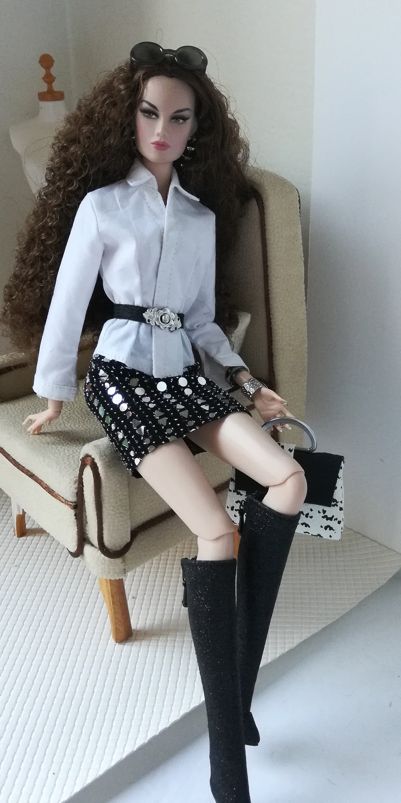 May include: A doll wearing a white button-down shirt, a black sequined skirt, and black boots. The doll is sitting on a beige armchair with a black and white handbag next to her.