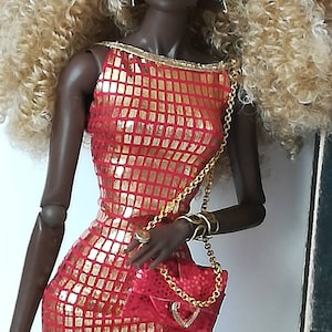 Pode incluir: Uma boneca de moda com cabelo loiro encaracolado usando um vestido de lantejoulas vermelho e dourado e uma bolsa vermelha com uma corrente dourada.