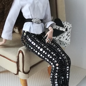 May include: A doll wearing a white button-down shirt with a black belt and a silver buckle. The doll is wearing black pants with silver sequins and black boots. The doll is holding a white and black handbag.