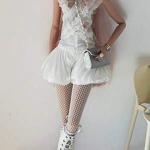 May include: A fashion doll wearing a white lace top, white shorts, white fishnet tights, and white lace-up boots. The doll has pink hair and is wearing sunglasses and pearl earrings. The doll is holding a gray clutch purse.