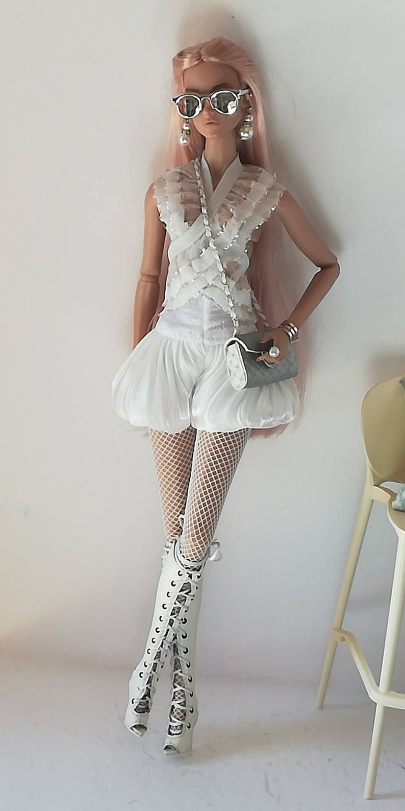 May include: A fashion doll wearing a white lace top, white shorts, white fishnet tights, and white lace-up boots. The doll has pink hair and is wearing sunglasses and pearl earrings. The doll is holding a gray clutch purse.