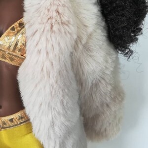 Puede incluir: Una mu&ntilde;eca con una chaqueta de piel sint&eacute;tica blanca sobre un top de lentejuelas doradas y leggings amarillos.