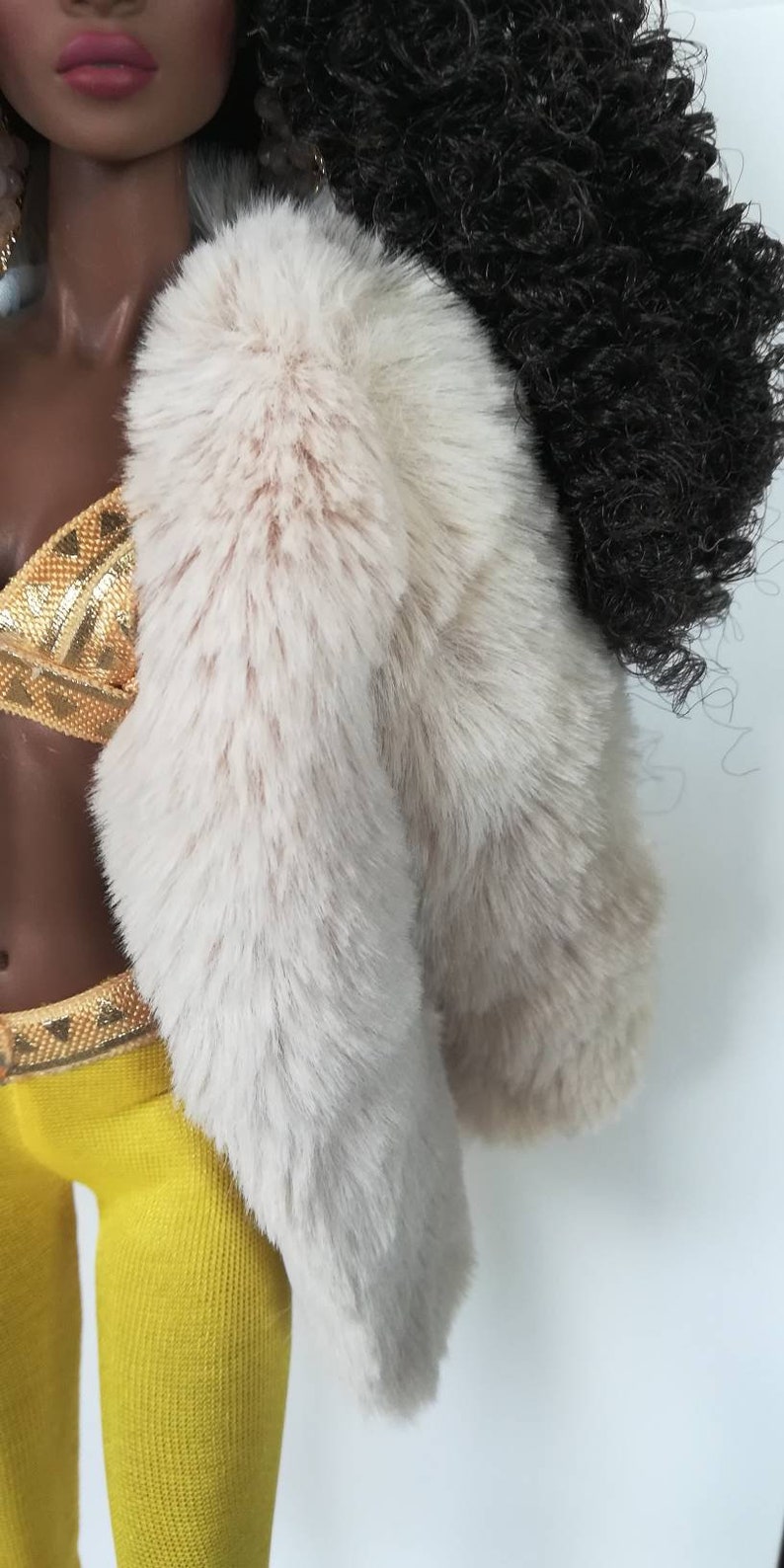 Puede incluir: Una mu&ntilde;eca con una chaqueta de piel sint&eacute;tica blanca sobre un top de lentejuelas doradas y leggings amarillos.
