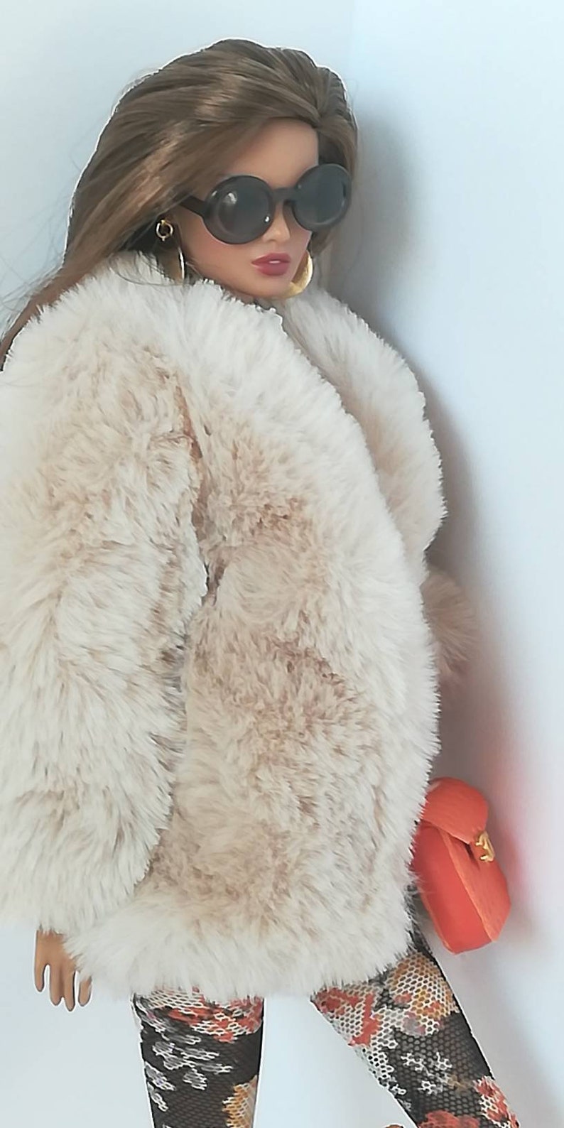 Puede incluir: Una mu&ntilde;eca con un abrigo de piel sint&eacute;tica de color crema, medias estampadas en negro y naranja y un peque&ntilde;o bolso de mano naranja. La mu&ntilde;eca lleva gafas de sol redondas grandes.