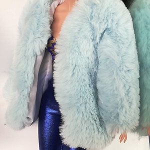 May include: A doll with long black hair wearing a light blue faux fur coat and blue metallic trousers. The doll has green eyeshadow and pink lipstick.