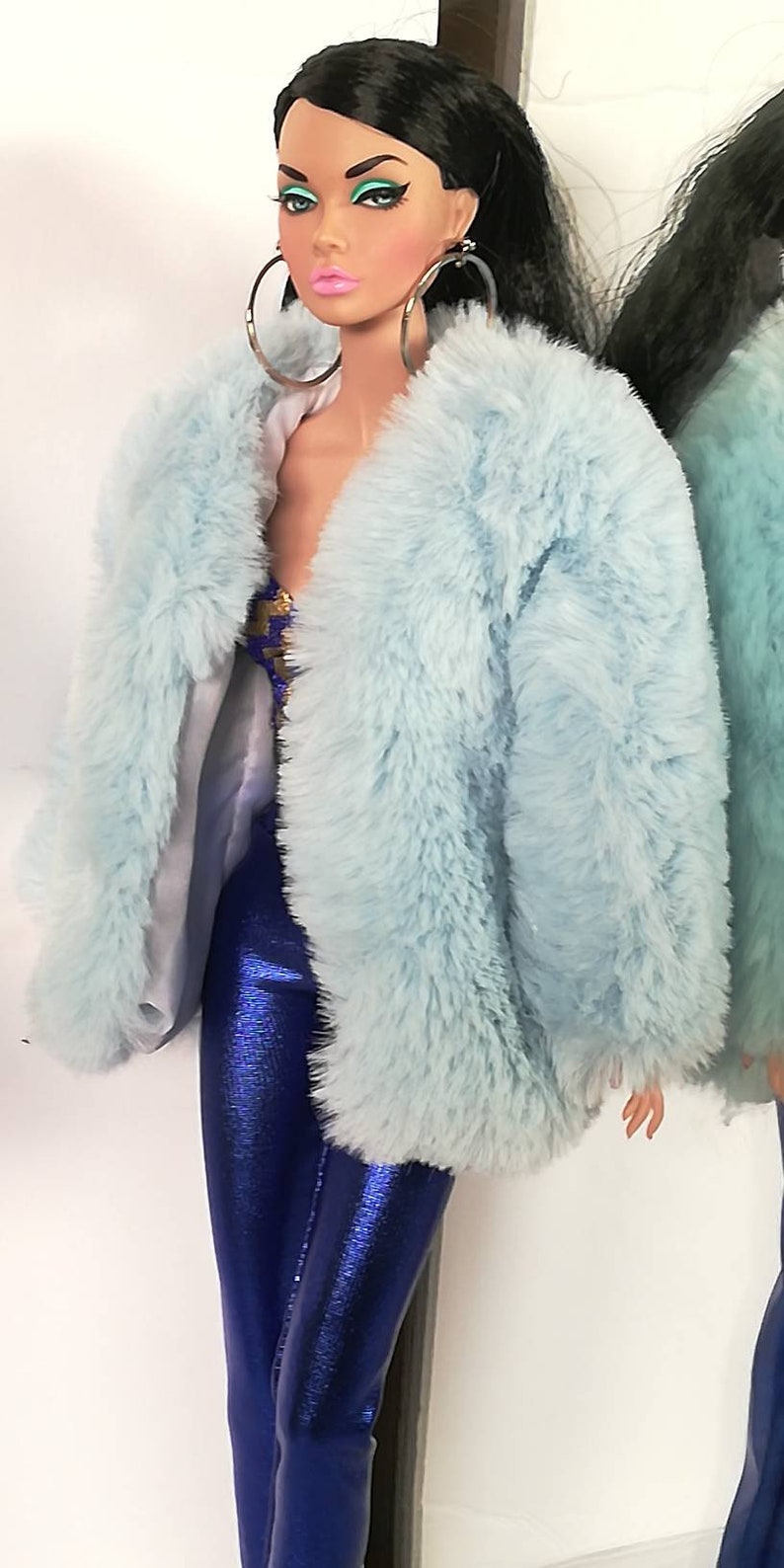 May include: A doll with long black hair wearing a light blue faux fur coat and blue metallic trousers. The doll has green eyeshadow and pink lipstick.