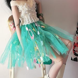 May include: A fashion doll in a cream top with pearl details and a turquoise tulle skirt with colourful ribbons. The doll wears sunglasses, earrings, and white boots with a wooden heel. The doll is seated on a white chair.