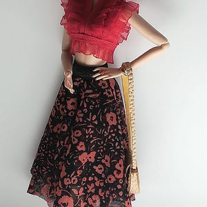 Op de afbeelding: Een modepop met een rood gerimpeld topje, een zwarte rok met rode bloemenprint, zwarte sandalen en een zonnebril. De pop houdt een bruine handtas vast.