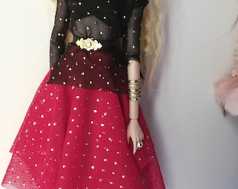 May include: A fashion doll wearing a black mesh top with gold dots, a red skirt with gold dots, and clear heels. The doll has blonde hair, sunglasses, and gold jewelry. The outfit includes a gold belt.