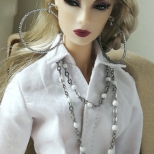 May include: A doll with blonde hair wearing a white button-down shirt with a silver chain necklace and a silver belt buckle. The doll is wearing silver hoop earrings.