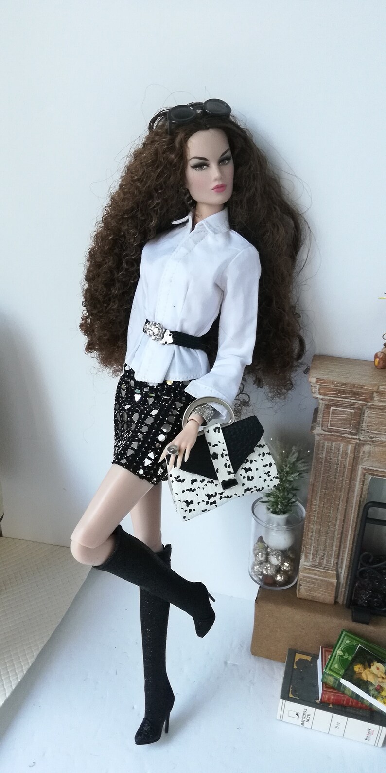May include: A doll wearing a white button-down shirt with a black belt, a black and white sequined skirt, black knee-high boots, and a black and white handbag. The doll has long brown curly hair and is wearing sunglasses.