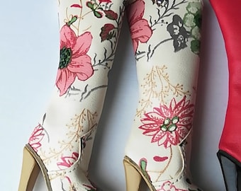 May include: A pair of white cowboy boots with a floral print. The boots have a high heel and a pointed toe. The floral print is in shades of pink, red, and green.