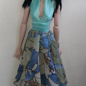 Könnte beinhalten: Eine Puppe trägt ein türkisfarbenes Halstuch und einen Rock mit blauem und grünem Blumendruck.