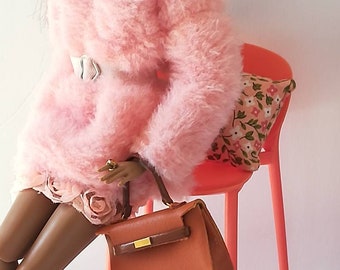 May include: A doll wearing a pink fuzzy sweater, a white belt, and pink boots. The doll is sitting on a bright orange stool with a floral patterned cushion. The doll is holding a brown leather handbag.