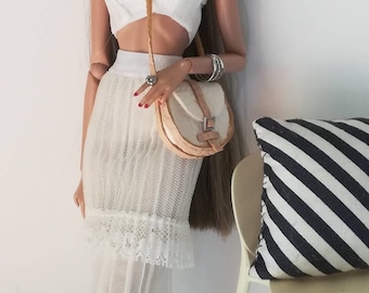 Può includere: Una bambola con un top bianco corto, una gonna bianca con bordo in pizzo e sandali bianchi. La bambola tiene una borsa a mano marrone chiaro con una fibbia argentata. La bambola ha i capelli lunghi e castani e indossa una fascia per capelli e orecchini argentati.