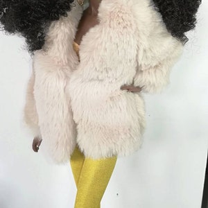 Puede incluir: Una mu&ntilde;eca con el pelo largo y negro rizado que lleva un abrigo de piel sint&eacute;tica blanco, leggings amarillos y botas negras.