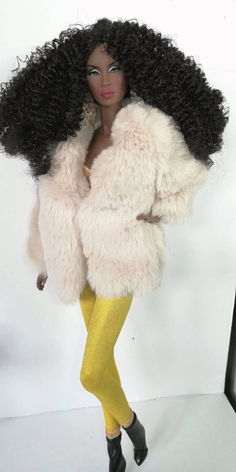 Puede incluir: Una mu&ntilde;eca con el pelo largo y negro rizado que lleva un abrigo de piel sint&eacute;tica blanco, leggings amarillos y botas negras.