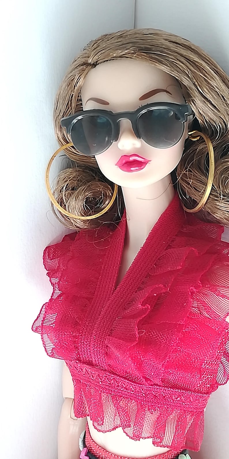 Op de afbeelding: Een pop met bruin haar die een rood kanten topje draagt, een zwarte zonnebril en gouden oorringen.