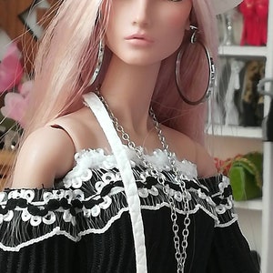 May include: A doll with pink hair wearing a white fedora, a black off-the-shoulder top with white lace trim, and silver hoop earrings. The doll is wearing a silver chain necklace and a white strap.