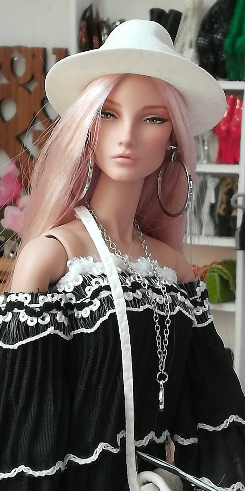 May include: A doll with pink hair wearing a white fedora, a black off-the-shoulder top with white lace trim, and silver hoop earrings. The doll is wearing a silver chain necklace and a white strap.