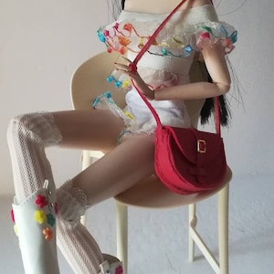 May include: A doll wearing a white dress with colorful embroidery, white boots with colorful flower embellishments, and a red handbag with a gold buckle. The doll has long black hair and is sitting on a white stool.
