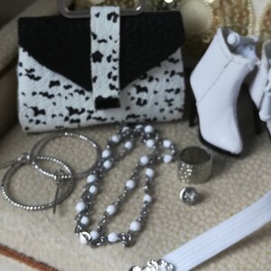 May include: A white and black patterned handbag with a silver clasp, a pair of white heeled boots, a silver chain necklace with white beads, and a silver ring on a white surface.