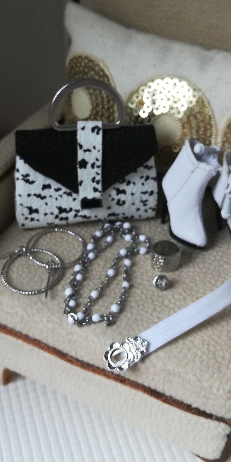 May include: A white and black patterned handbag with a silver clasp, a pair of white heeled boots, a silver chain necklace with white beads, and a silver ring on a white surface.