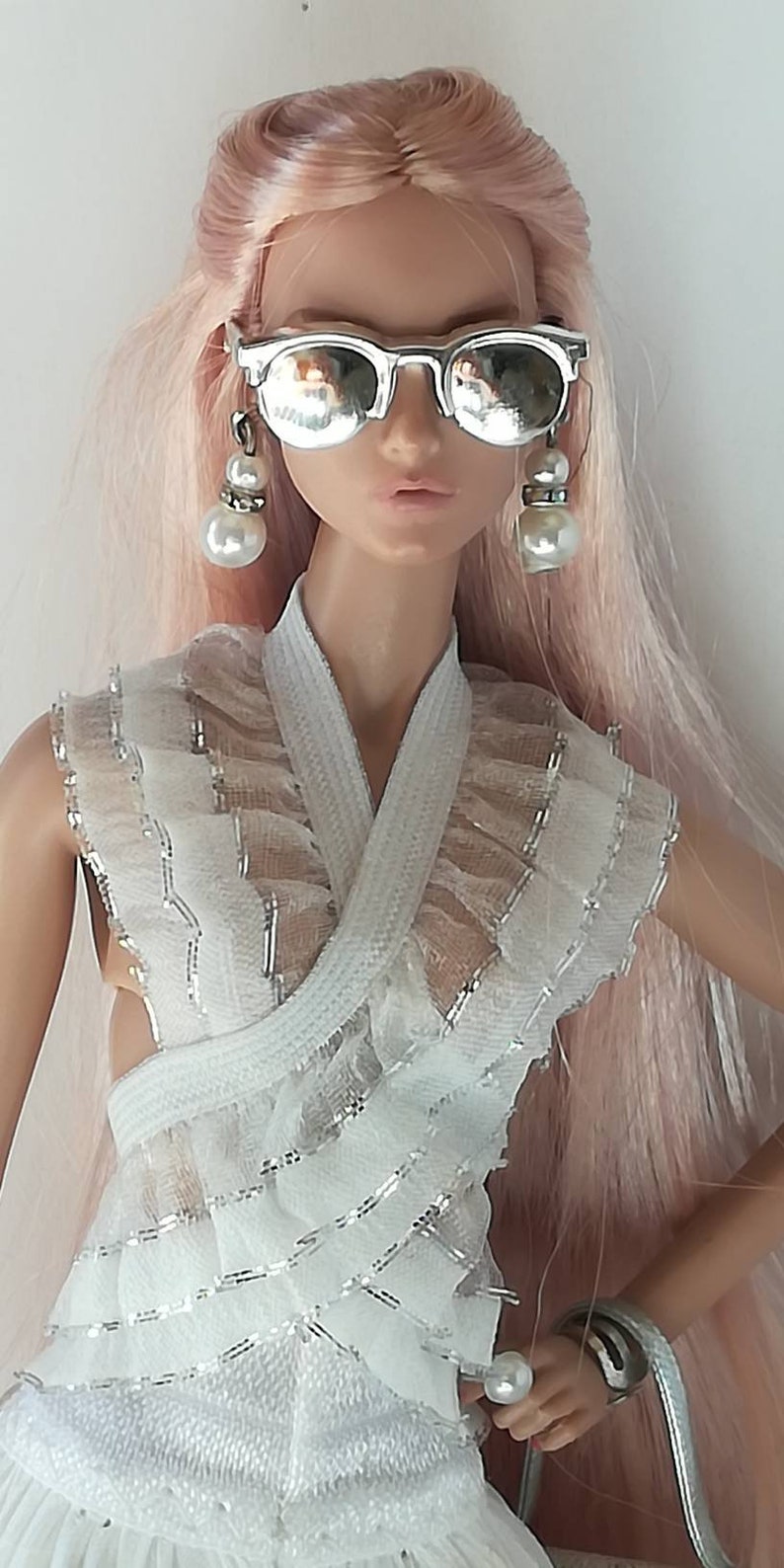 May include: A doll with long pink hair wearing a white ruffled dress, silver mirrored sunglasses, and pearl earrings.