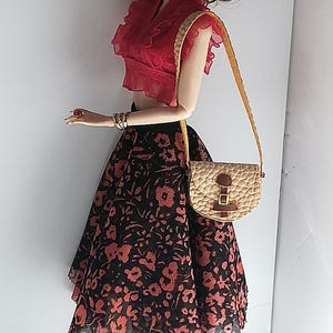 Op de afbeelding: Een modepop met lang bruin haar, gekleed in een rood topje, een zwarte en rode bloemenrok, gouden oorringen en zwarte plateau sandalen. De pop houdt een bruin en beige handtas vast met een gouden band.