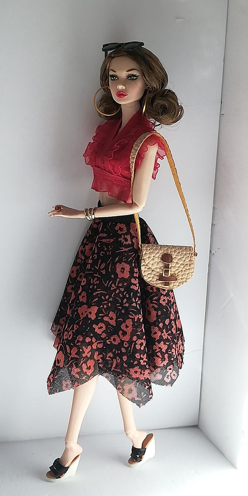 Op de afbeelding: Een modepop met lang bruin haar, gekleed in een rood topje, een zwarte en rode bloemenrok, gouden oorringen en zwarte plateau sandalen. De pop houdt een bruin en beige handtas vast met een gouden band.