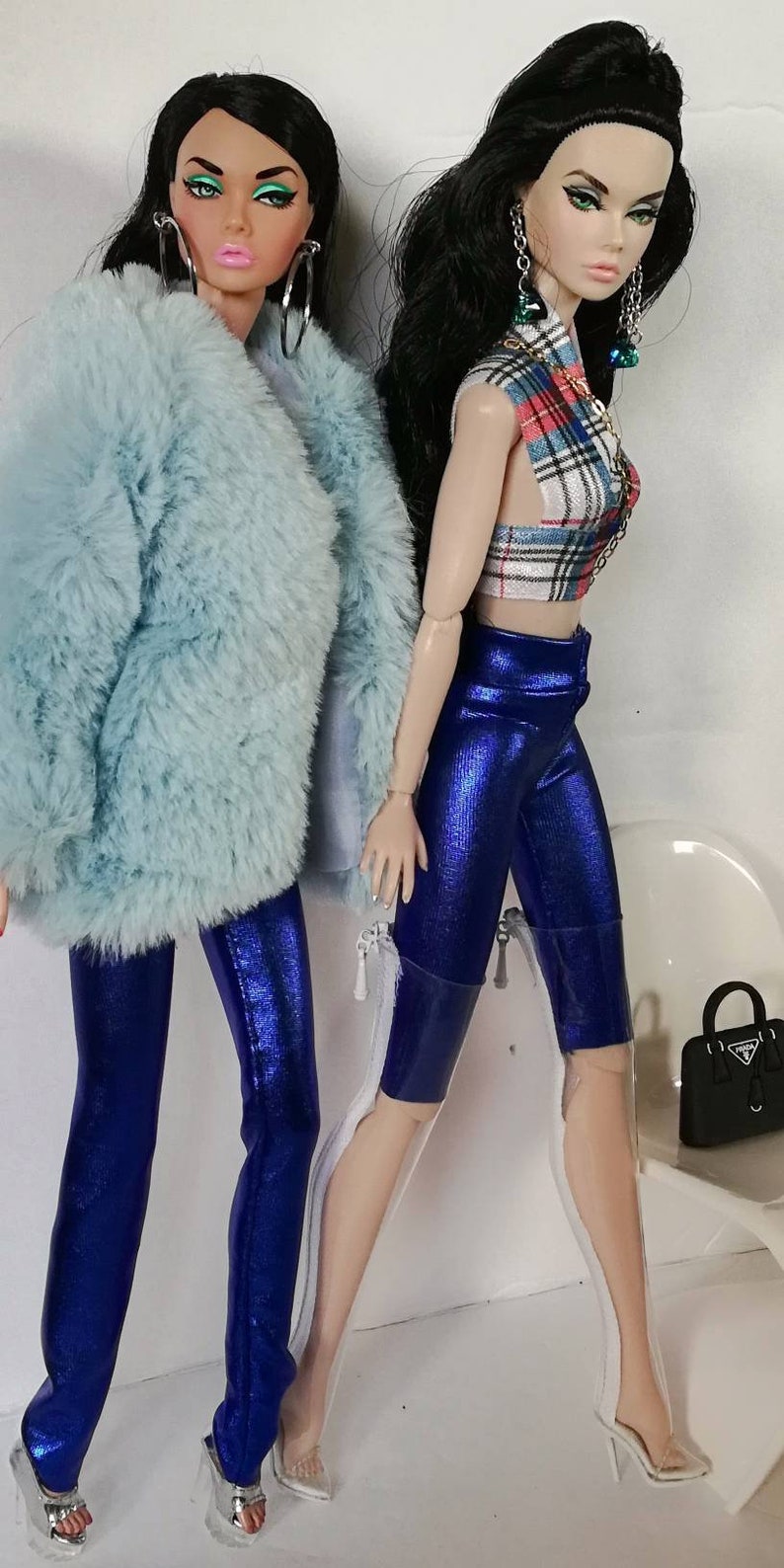 May include: Two fashion dolls wearing blue trousers and white platform heels. One doll is wearing a light blue fluffy jacket and the other doll is wearing a red and white checked top.