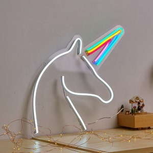 May include: White neon unicorn head sign with a rainbow horn. The sign is mounted on a wall with a string of fairy lights in the foreground.