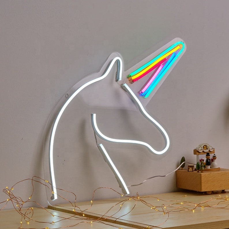 May include: White neon unicorn head sign with a rainbow horn. The sign is mounted on a wall with a string of fairy lights in the foreground.