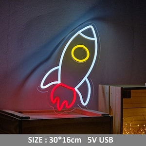 May include: White neon rocket ship sign with a yellow circle in the middle and a red flame. The sign is 30 inches by 16 inches and powered by a 5V USB.