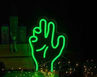 May include: Green neon sign shaped like a hand with three fingers raised. The sign is on a dark background with a string of fairy lights in the foreground. The sign reads "SIZE: 30*21cm/40*28cm/50*35cm".
