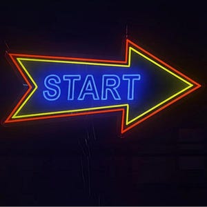 May include: Neon sign in the shape of an arrow, with the word "START" in blue neon. The arrow has a yellow outline with a red border, all against a dark background. The sign is designed to point to the right.