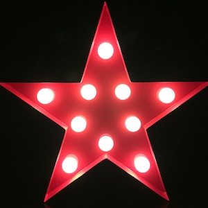 Puede incluir: Una estrella roja con diez bombillas blancas. La estrella es de color rojo brillante con un acabado brillante. Las bombillas están espaciadas uniformemente y emiten un brillo cálido. El fondo es negro.