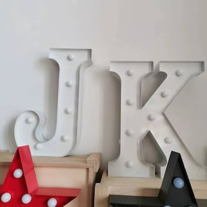 以下が含まれることがあります： 木製の棚の上に、電球が付いた白い金属製の文字JとKが置かれています。手前に、電球が付いた赤い星形と黒い星形のライトがあります。