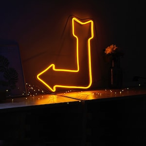 May include: A neon yellow arrow sign with a 90-degree bend, illuminated against a dark background. The sign is mounted on a wall and is surrounded by string lights.