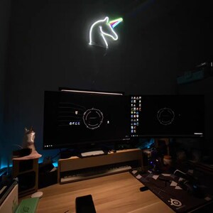 May include: A dark room with a desk and two computer monitors. The monitors are displaying a dark background with a circular design. A neon unicorn light is hanging on the wall above the monitors. There is a small wooden shelf with a white figurine on it.