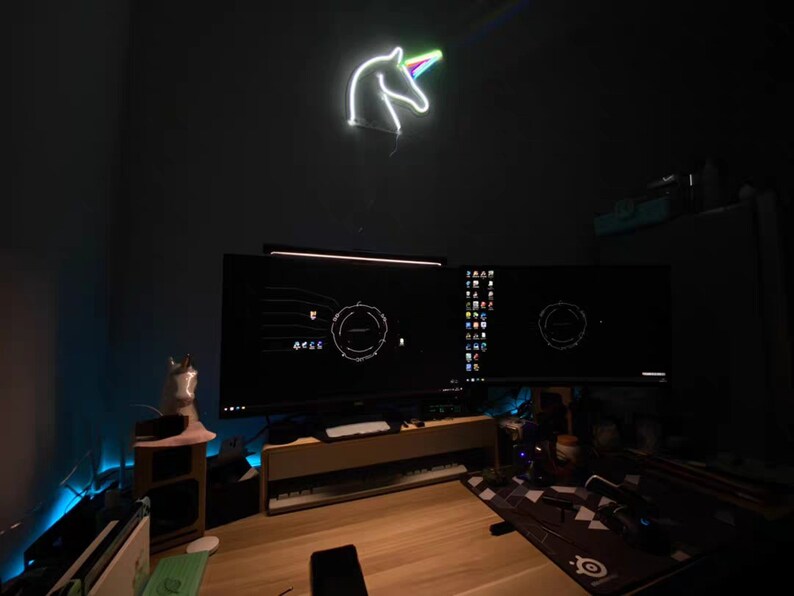 May include: A dark room with a desk and two computer monitors. The monitors are displaying a dark background with a circular design. A neon unicorn light is hanging on the wall above the monitors. There is a small wooden shelf with a white figurine on it.