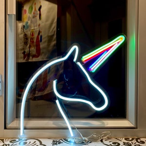 May include: A white neon unicorn head sign with a rainbow horn. The sign is lit up and is sitting on a window sill.