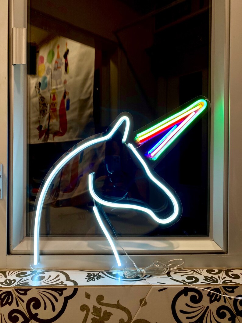 May include: A white neon unicorn head sign with a rainbow horn. The sign is lit up and is sitting on a window sill.