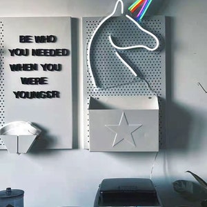 May include: A white pegboard with a rainbow unicorn neon sign, a white metal shelf with a star cutout, and a motivational quote that reads "Be who you needed when you were younger".