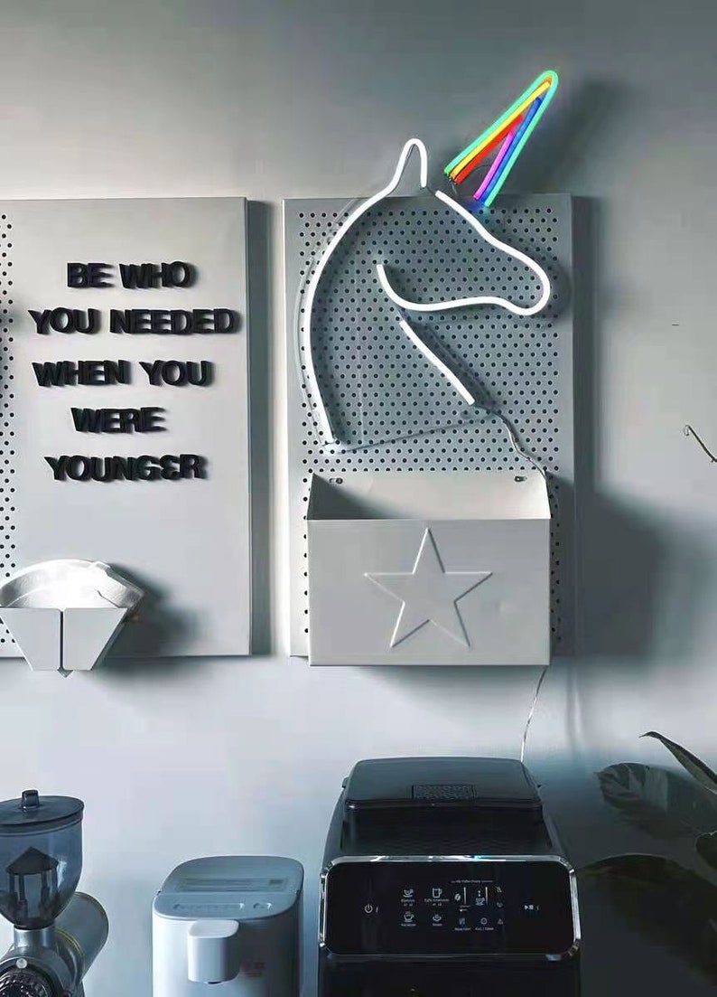 May include: A white pegboard with a rainbow unicorn neon sign, a white metal shelf with a star cutout, and a motivational quote that reads "Be who you needed when you were younger".