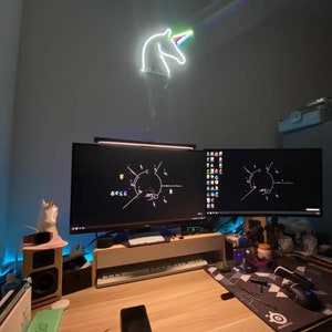 May include: A desk with two computer monitors, a keyboard, a mouse, and a unicorn neon sign on the wall. The desk is made of wood and has a light wood finish. The monitors are both black and have a dark screen. The keyboard is black and has a white backlight. The mouse is black and has a white scroll wheel. The unicorn neon sign is white and has a rainbow mane and horn.