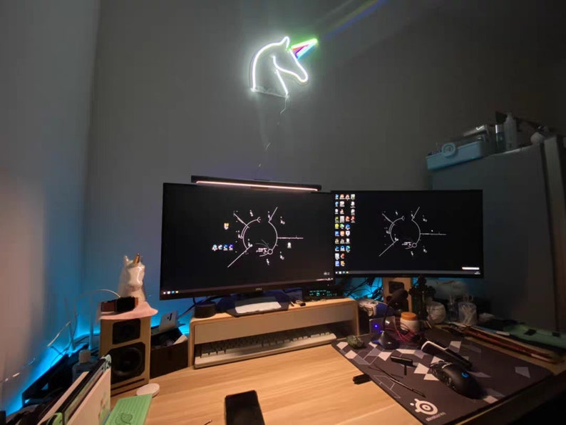 May include: A desk with two computer monitors, a keyboard, a mouse, and a unicorn neon sign on the wall. The desk is made of wood and has a light wood finish. The monitors are both black and have a dark screen. The keyboard is black and has a white backlight. The mouse is black and has a white scroll wheel. The unicorn neon sign is white and has a rainbow mane and horn.