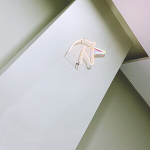May include: A white wall with a neon unicorn head sign with a rainbow horn. The sign is mounted on the wall and is lit up.