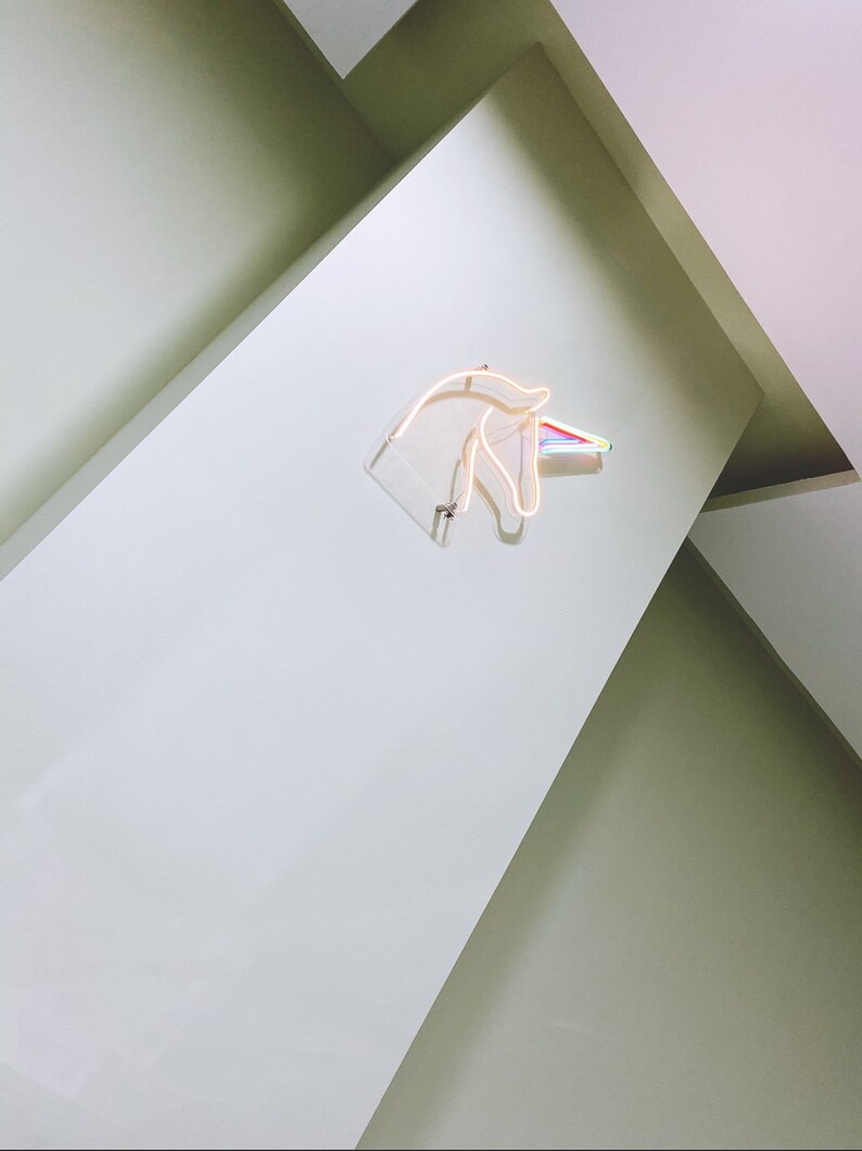 May include: A white wall with a neon unicorn head sign with a rainbow horn. The sign is mounted on the wall and is lit up.