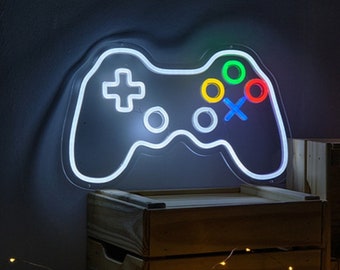 May include: A neon sign in the shape of a video game controller, with a white outline and colorful buttons. The controller measures 39 cm x 24 cm. The sign is lit up and mounted on a dark surface.